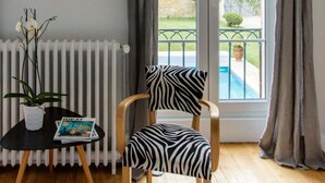 A room with a wooden floor, a zebra-patterned chair, a small table with a magazine, a potted plant, a radiator, and a window with curtains.