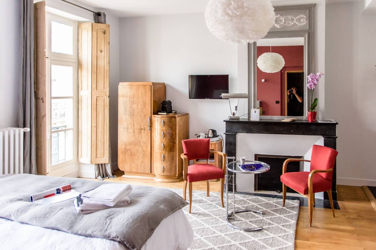 A hotel room with a bed, a wooden wardrobe, a red chair, a small table, a fireplace, and a TV mounted on the wall.