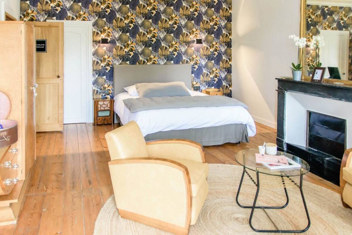 A bedroom with a bed, two chairs, a glass coffee table, a fireplace, and a floral wallpaper.
