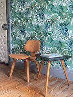 A room with a wooden chair and a small table, a tufted headboard, and a wall with a tropical leaf pattern.