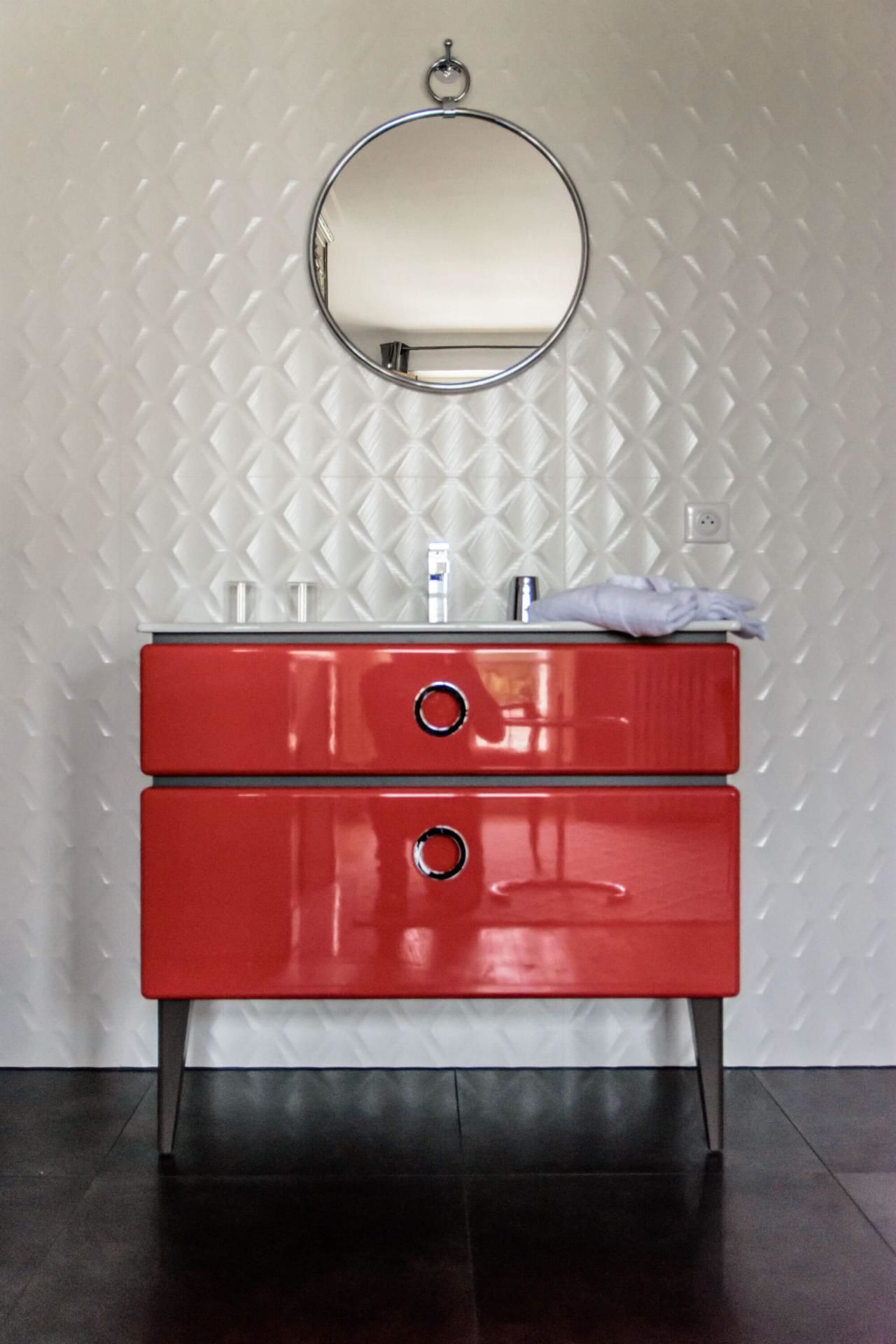 Une coiffeuse rouge surmontée d’un miroir rond, le tout placé devant un mur à texture.
