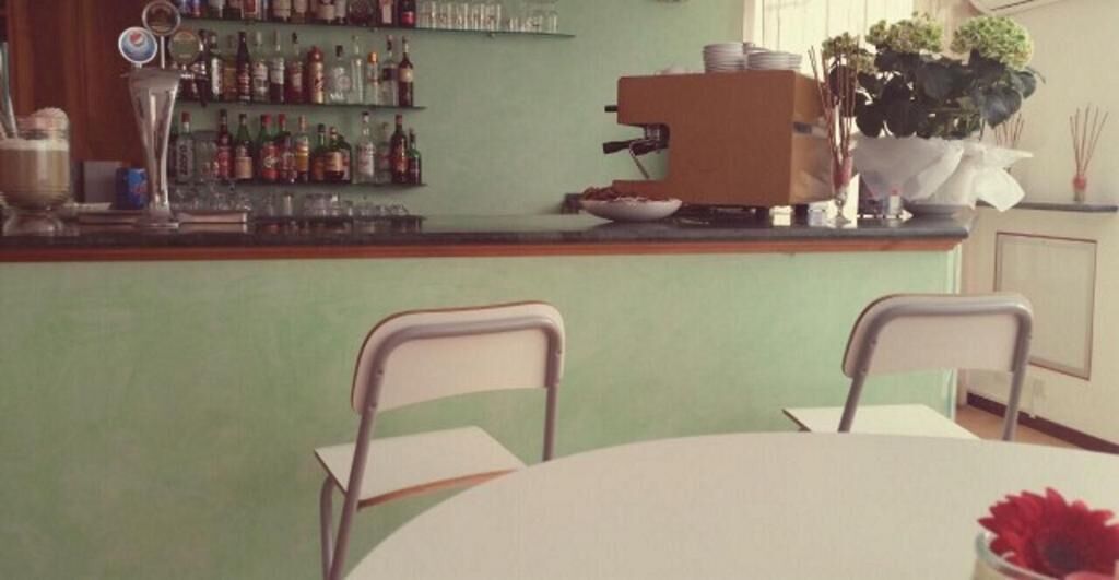 Een bar met een toonbank, planken vol flessen, een koffieapparaat en stoelen.