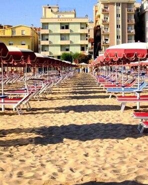 A beach with rows of sun loungers under red umbrellas, a building labeled "AUGUSTA" in the background, and sandy ground. - Hotel Augusta (Gabicce Mare)