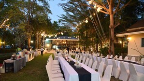 Een evenementenruimte buiten, met lange tafels klaar voor een diner, omringd door bomen en lichtslingers.