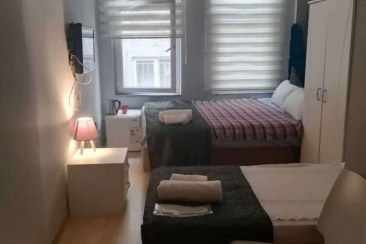 A small, well-lit room with two beds, a wardrobe, a desk with a lamp, and an air conditioning unit.