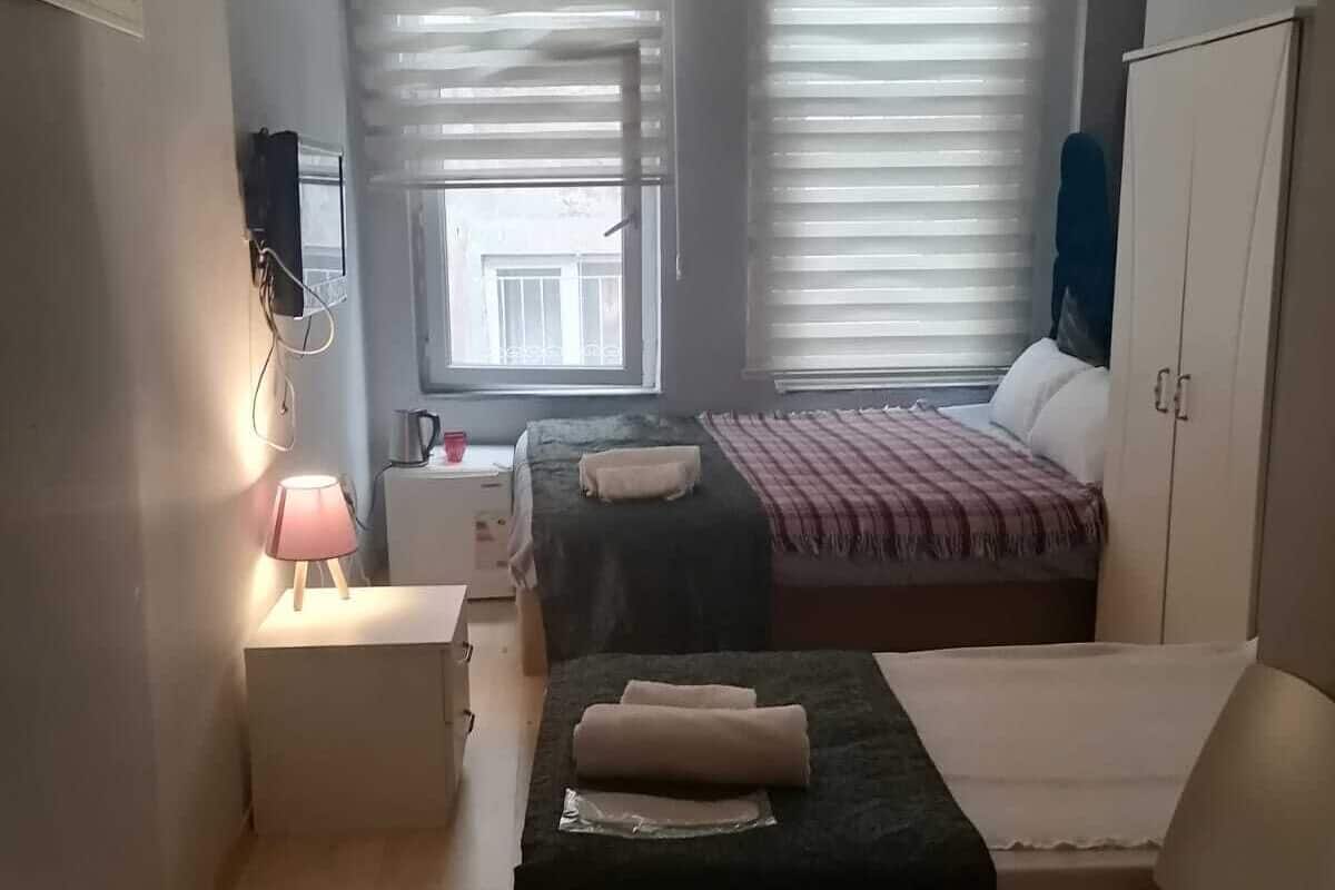 A small, well-lit room with two beds, a desk, a chair, and an air conditioning unit.
