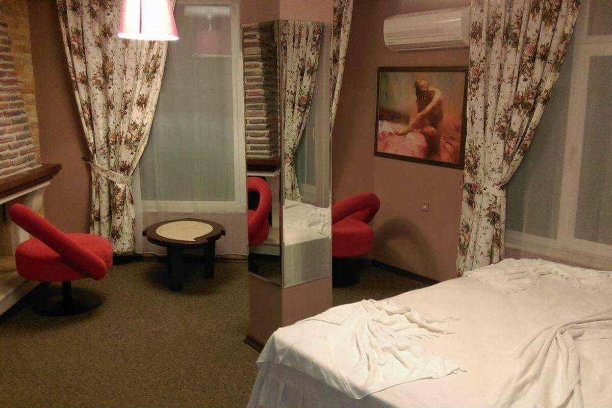Quarto de hotel com uma cama, duas poltronas vermelhas, uma mesa redonda pequena e um espelho.
