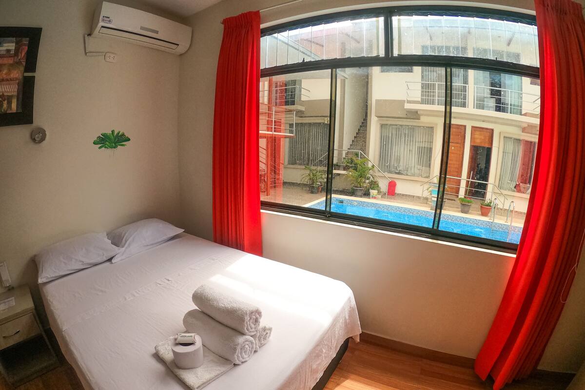 A hotel room with a bed, a window with red curtains, and a view of a swimming pool.
