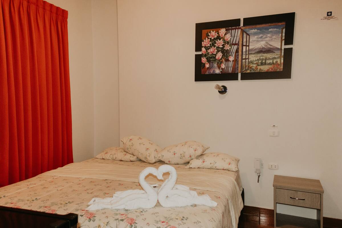 A hotel room with a bed, floral bedding, a heart-shaped towel arrangement, a nightstand, and a wall-mounted picture.