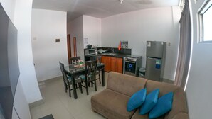 A modern living room with a brown sofa and blue cushions, a dining area with a glass table and chairs, and a kitchen with appliances and a window. - Residencial Marina House (La Banda de Shilcayo)