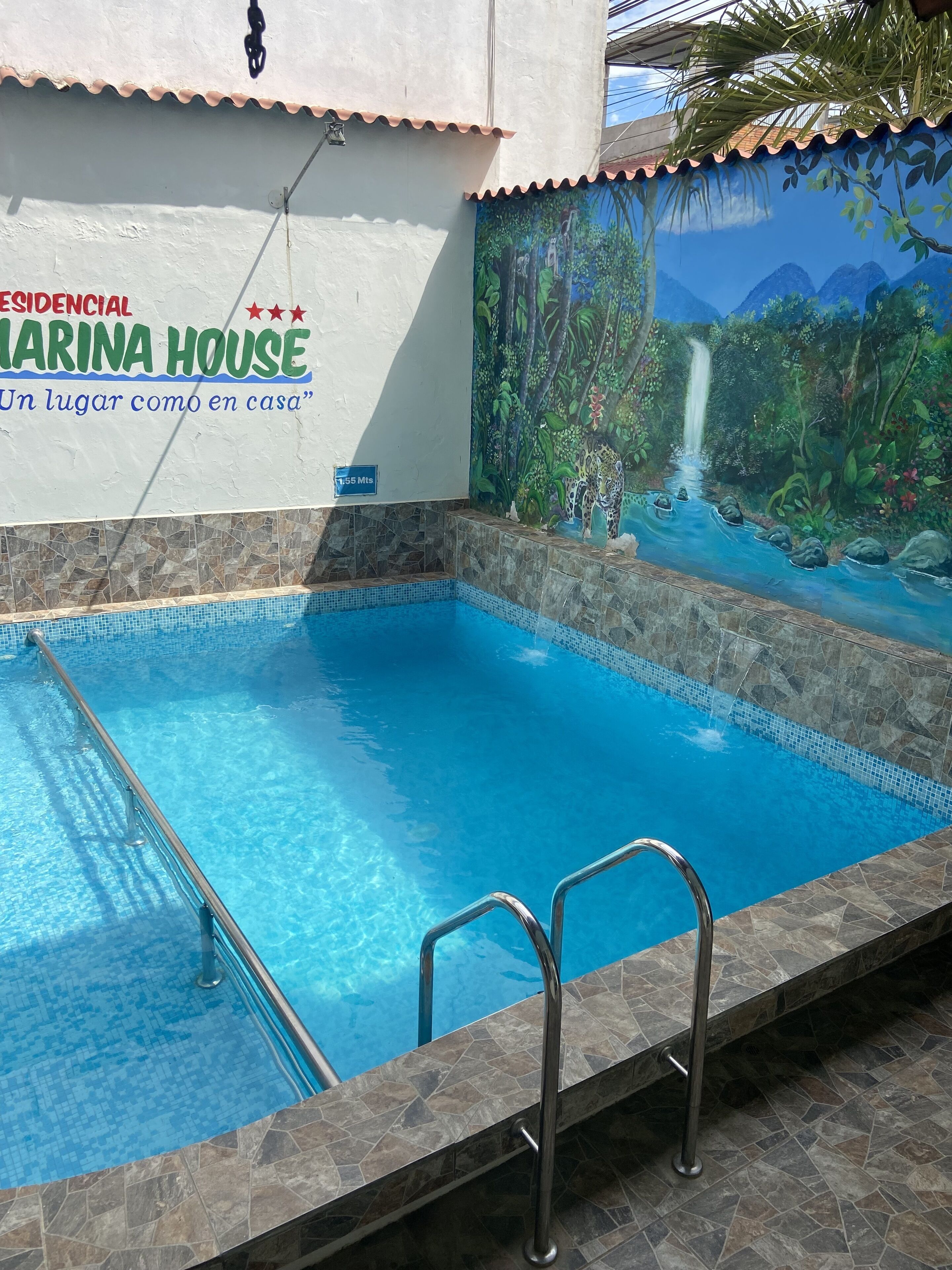 Una piscina con un mural de una selva y una cascada, un letrero que dice "MARINA HOUSE Un lugar como en casa", y un indicador de distancia de 1,55 Mts.
