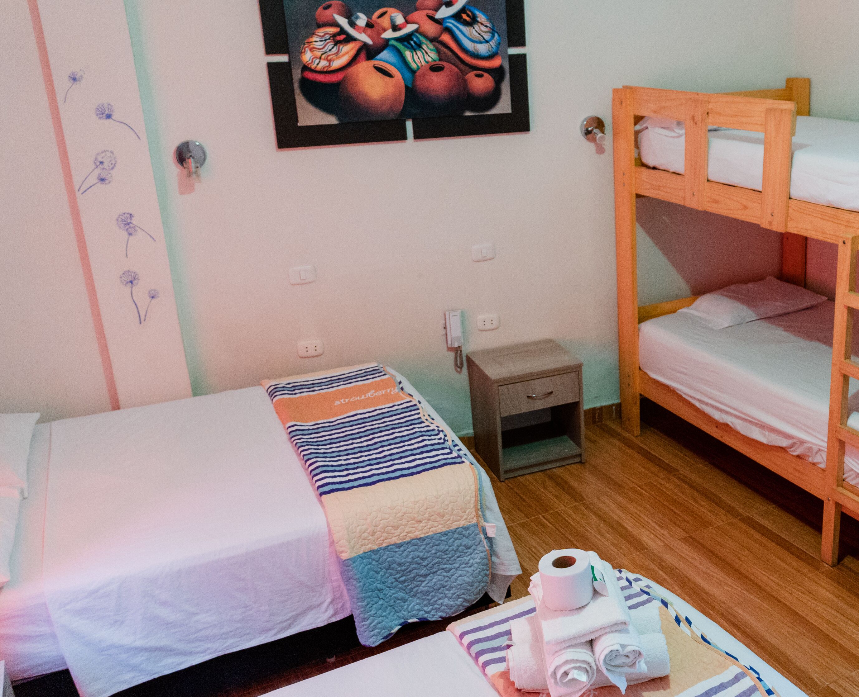 Quarto com duas camas de solteiro, uma cama beliche de madeira, uma pequena mesa de cabeceira com luminária e uma parede com um quadro colorido.