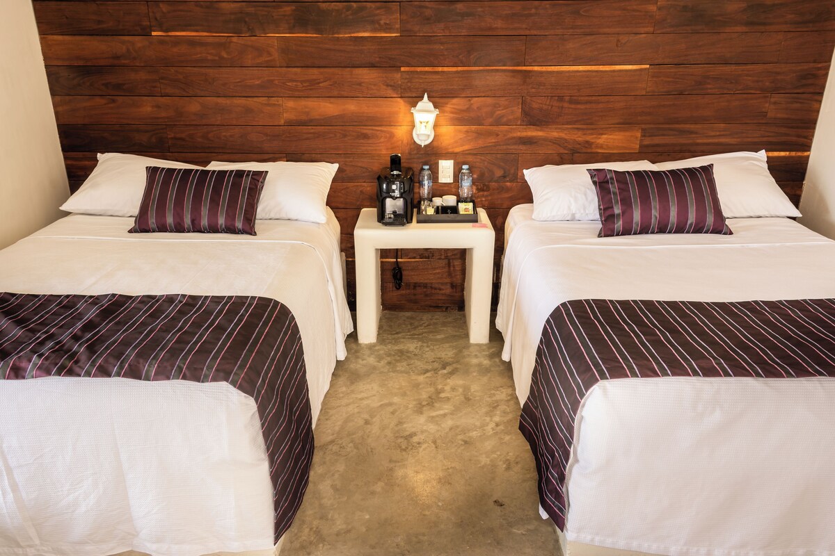 Una habitación de hotel con dos camas, una mesita de noche con cafetera y un cabecero de madera.