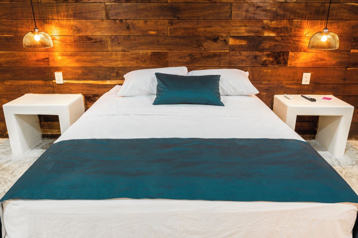 Una cama con sábanas blancas y un plaid color turquesa, dos mesitas de noche con lámparas, y un cabecero de madera.