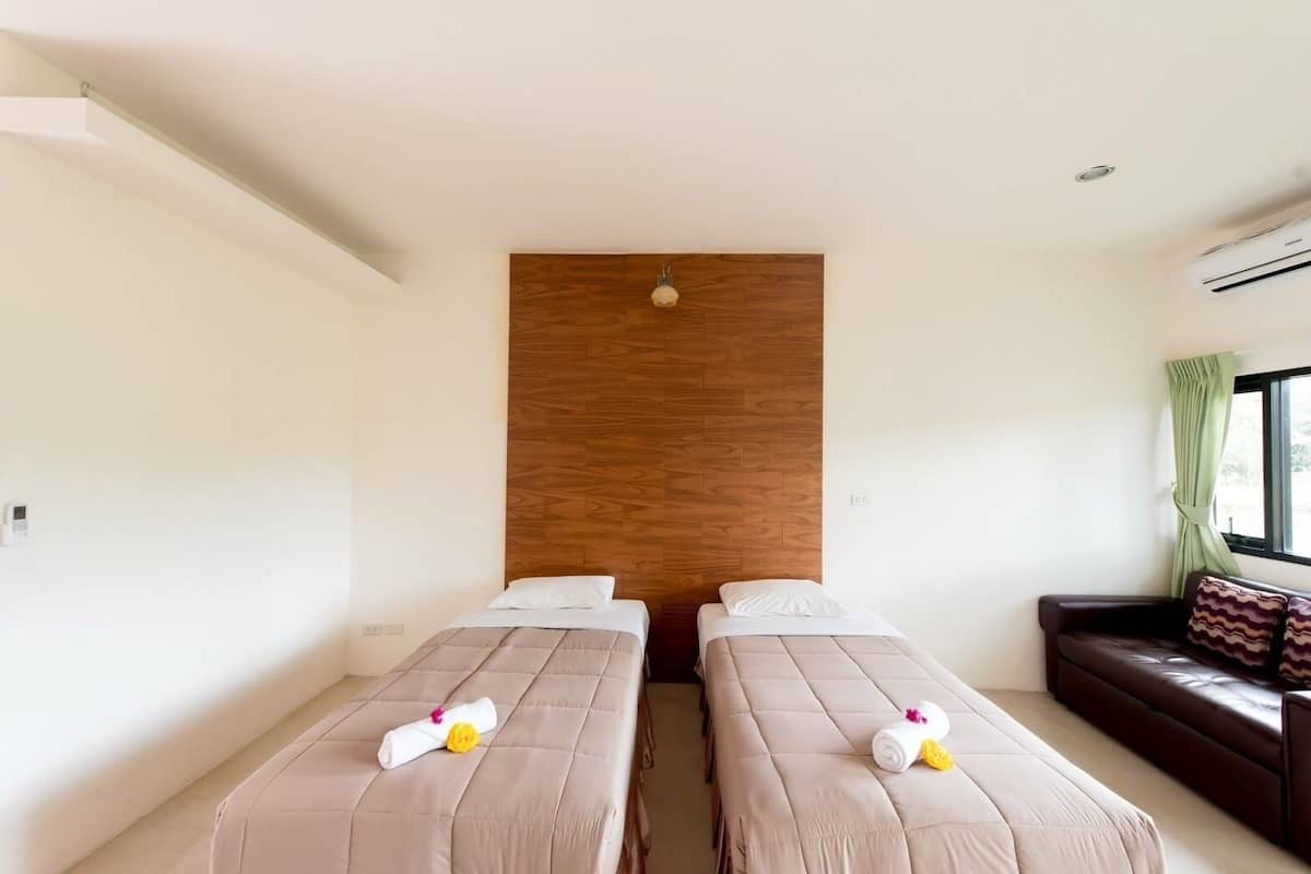 A hotel room with two beds, a wooden accent wall, a window with curtains, and a leather sofa.