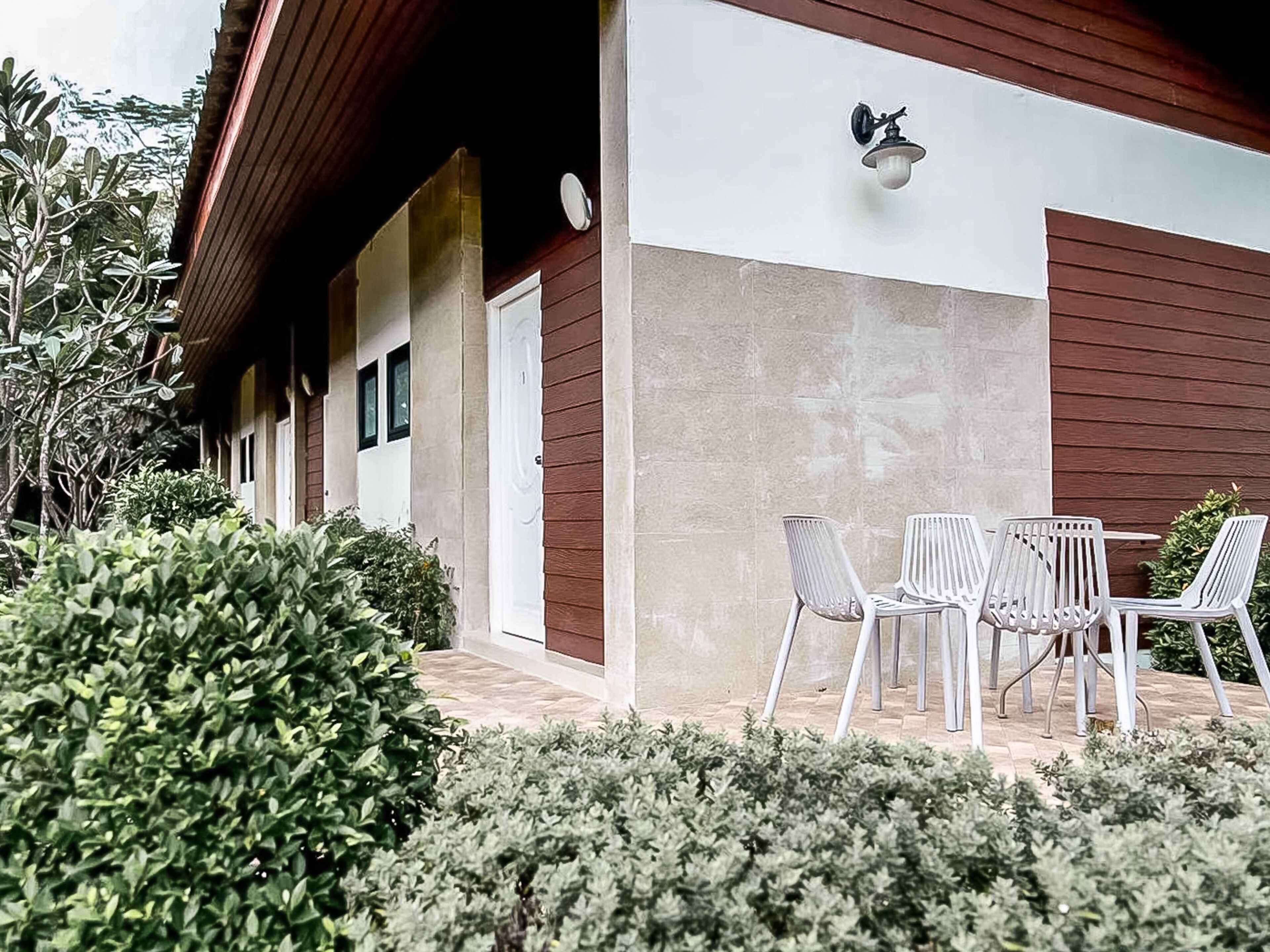 A building with a modern design, featuring a combination of wooden and stone exterior walls. There is a patio area with white metal chairs and a table, surrounded by well-maintained greenery.