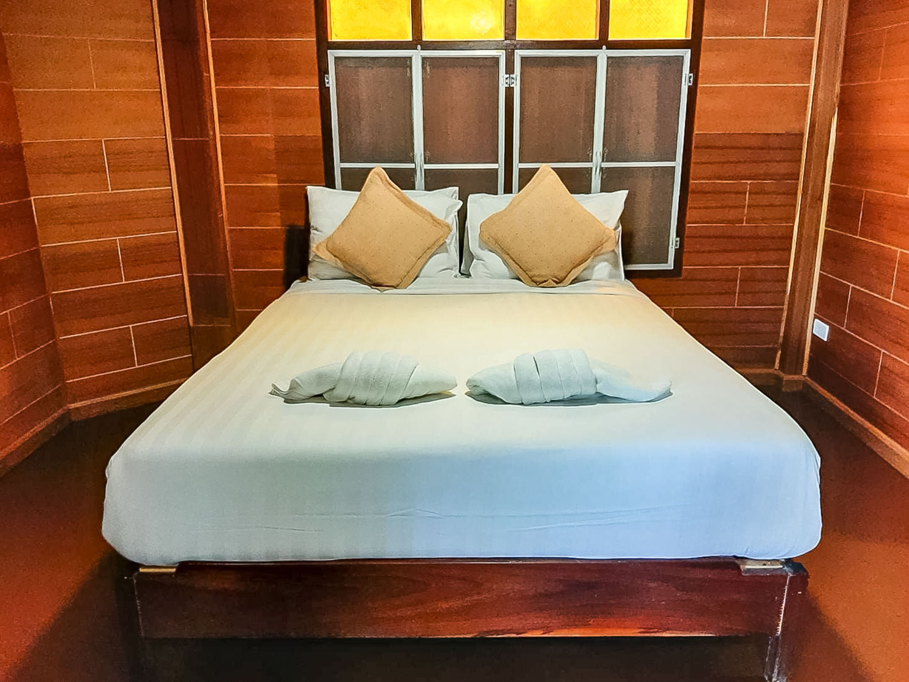A neatly made bed with two pillows and two folded towels in a room with brick walls and a window with blinds.