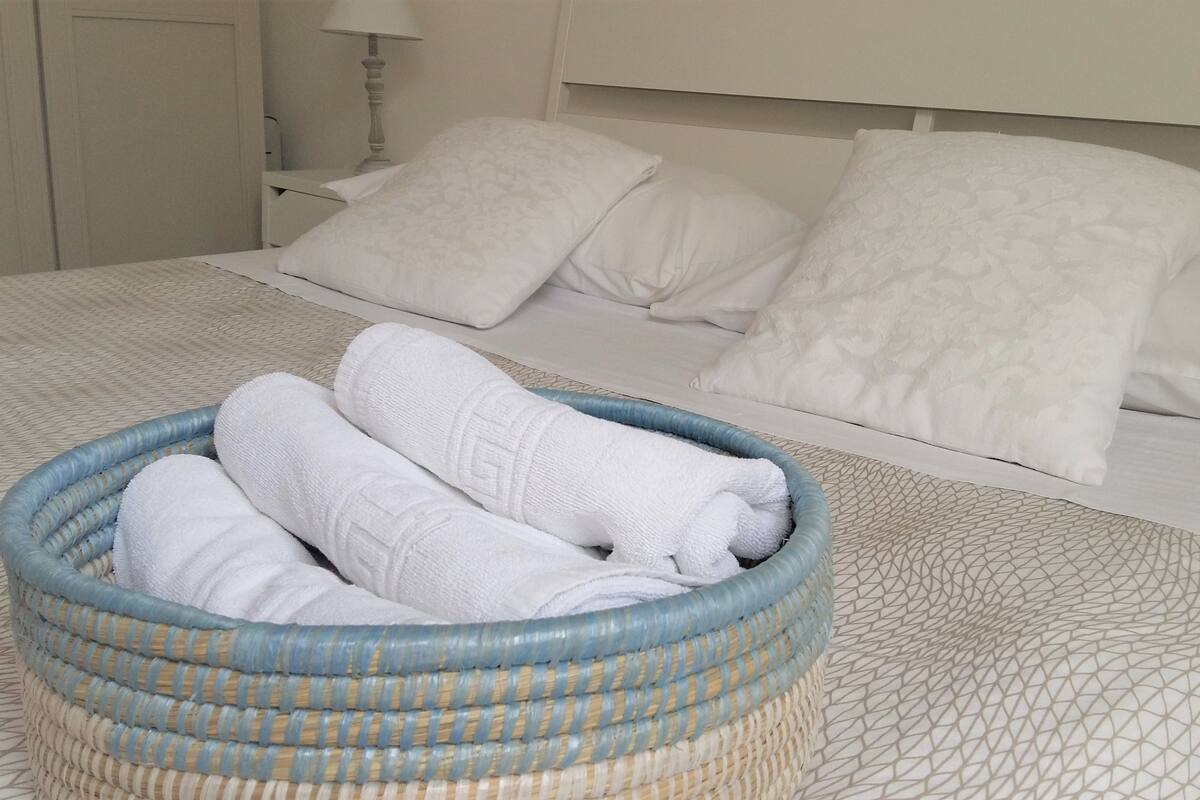 A neatly made bed with white linens and pillows, a blue woven basket with folded towels, and a patterned throw on the bed.