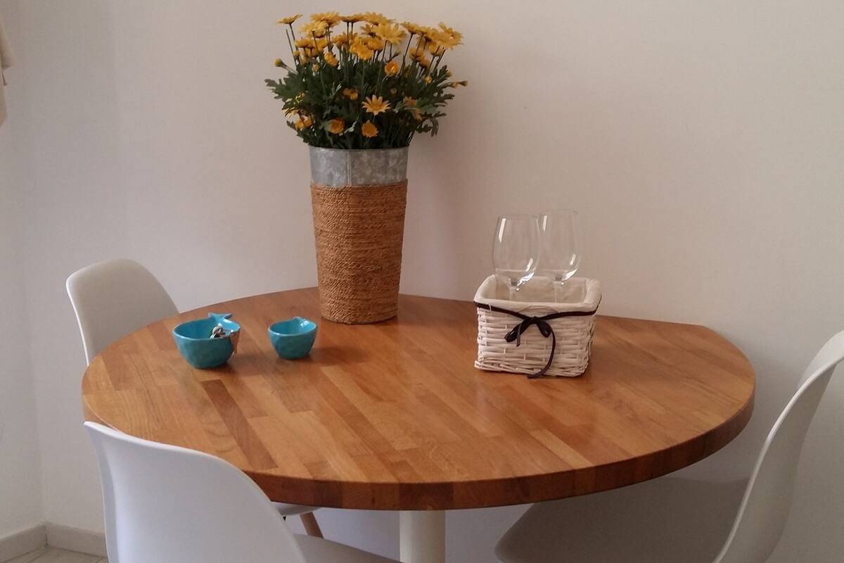 A round wooden dining table with two white chairs, a woven basket with two glasses, and a vase with yellow flowers.