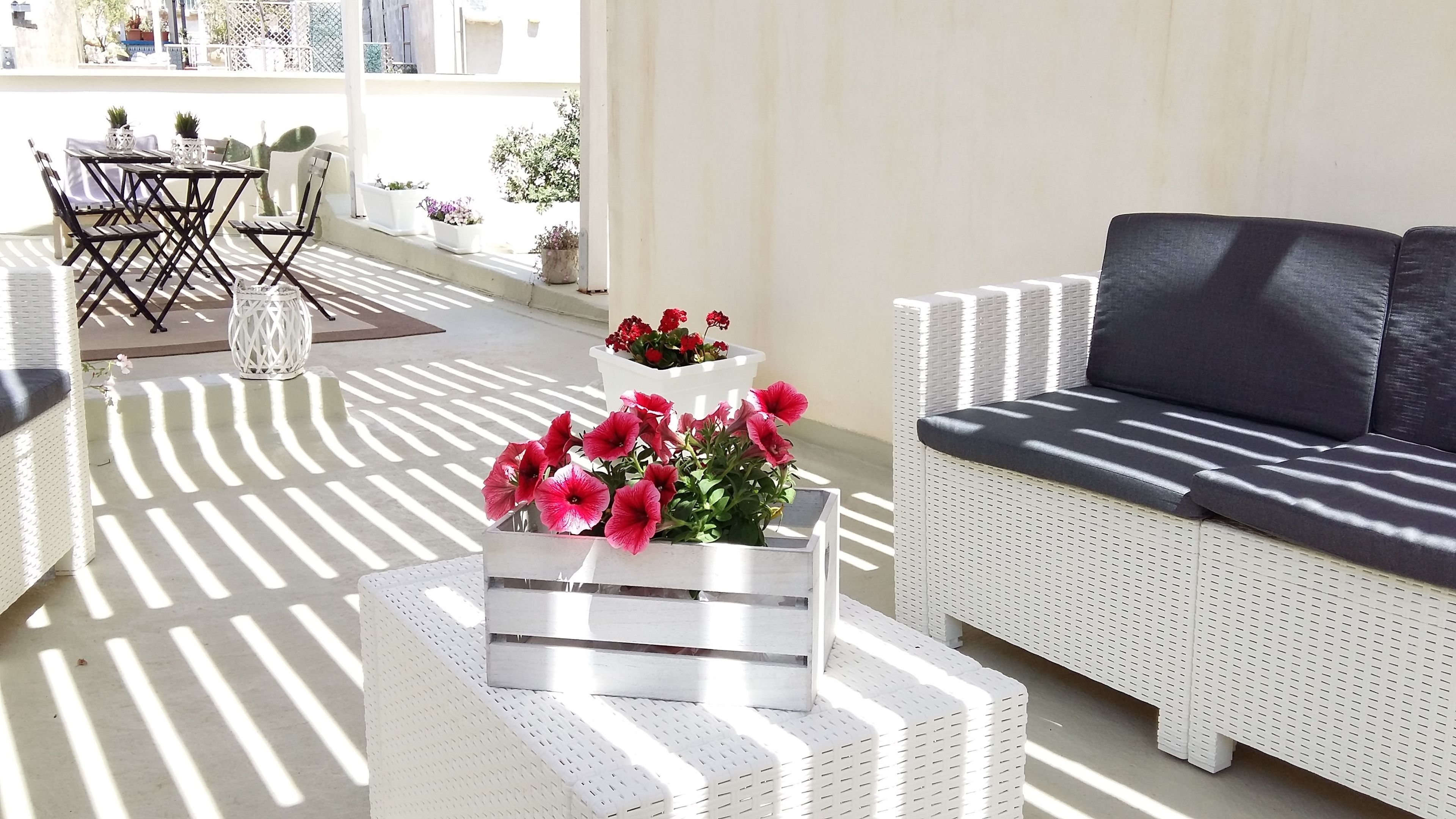 A sunlit patio area with a white wicker sofa, a small white table with a vase of pink flowers, and a white wicker chair.