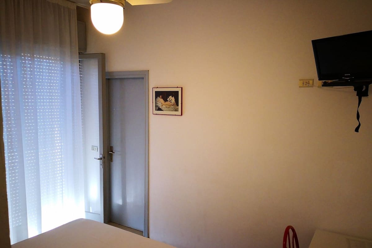 Un dormitorio con cama, una puerta, televisor montado en la pared y ventana con cortinas.