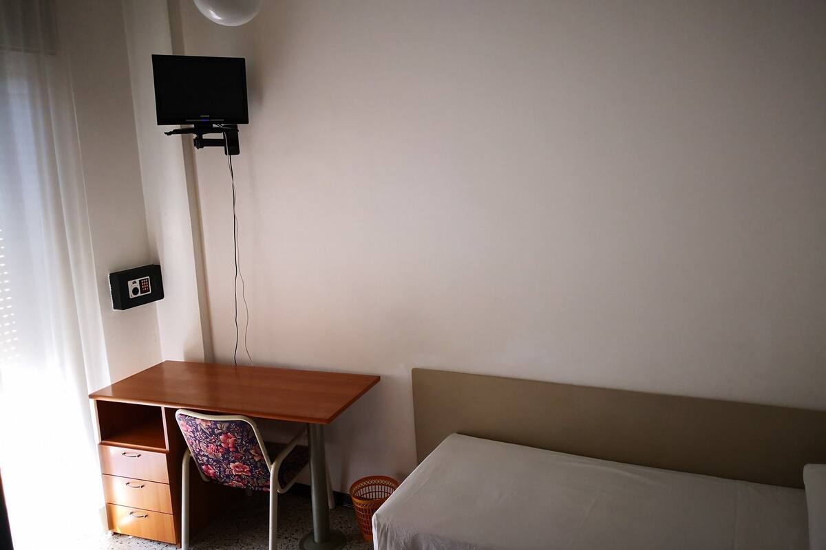 Una habitación con cama, escritorio, silla, televisor montado en la pared y ventilador de techo.