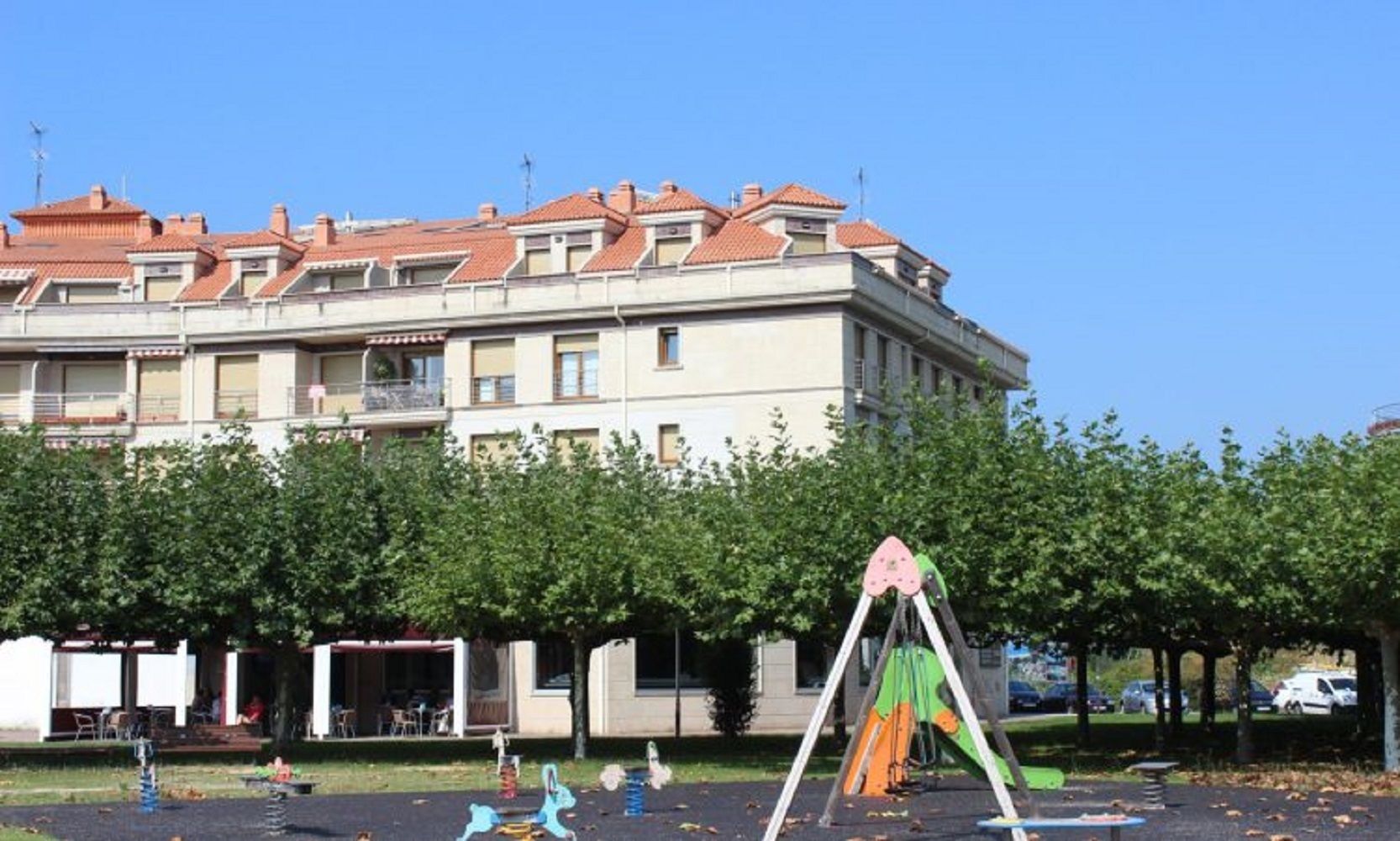 Un parque infantil con columpios y una estructura para escalar frente a un edificio con varios balcones y techo de tejas rojas.