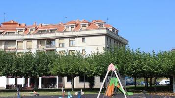 Un parque infantil con columpios y una estructura para escalar frente a un edificio con varios balcones y techo de tejas rojas.
