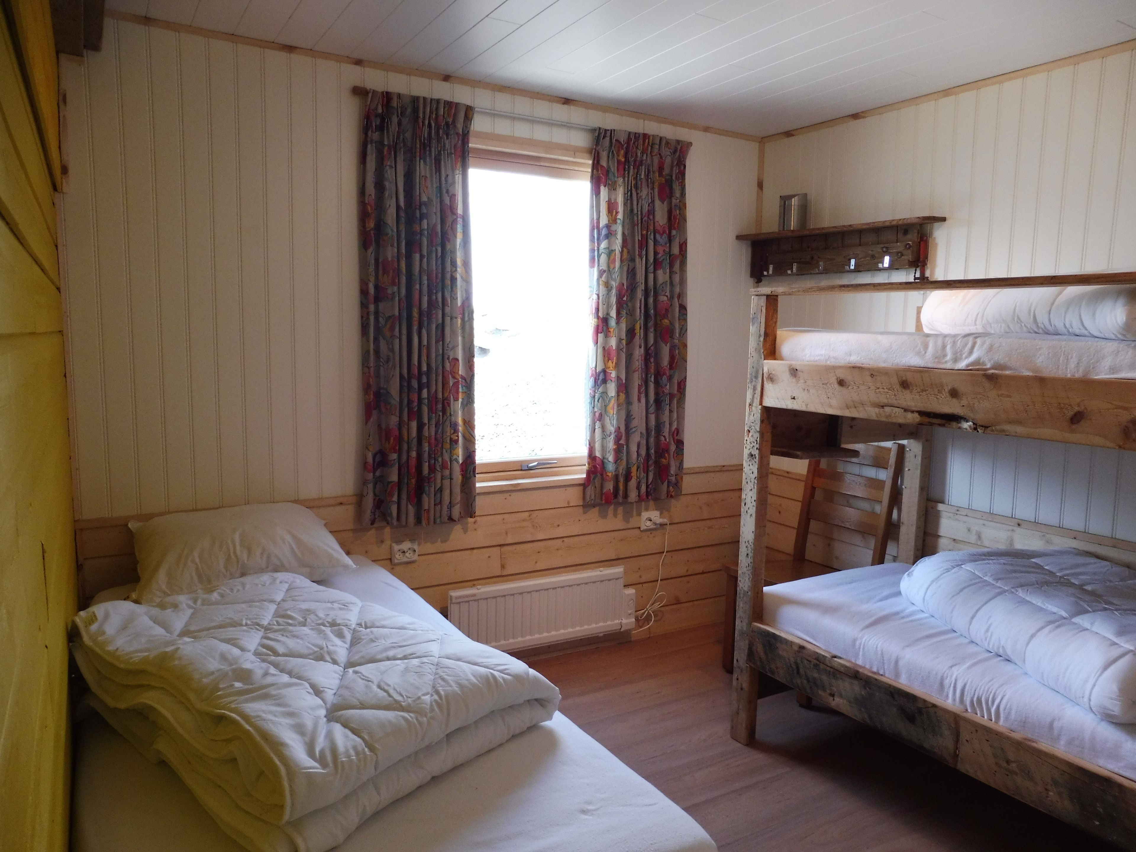 A room with a wooden bunk bed, a single bed, a window with curtains, and a radiator.