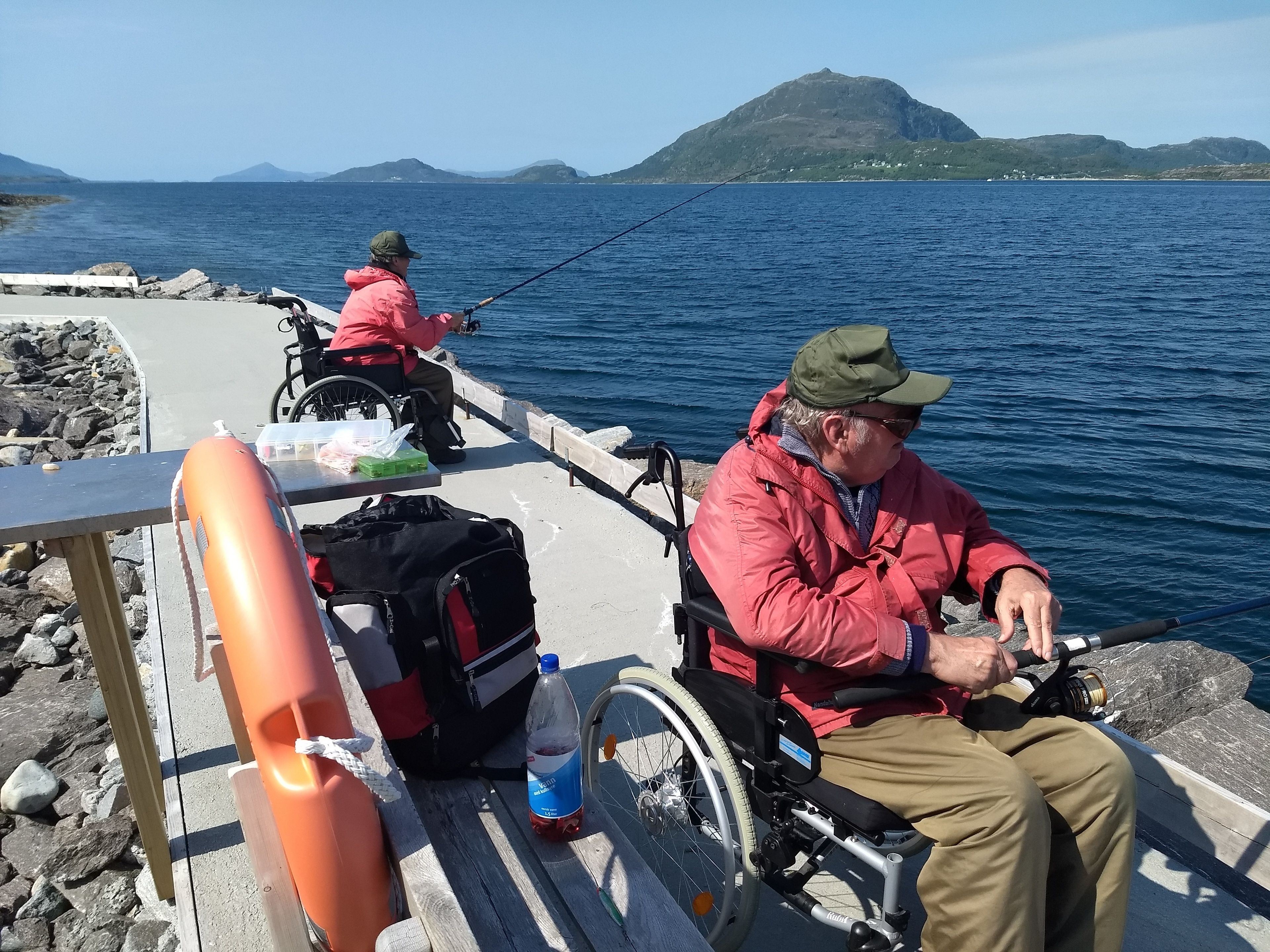 Two individuals in wheelchairs fishing by a rocky shoreline with a clear view of the sea and distant mountains.