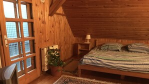 Ein gemütliches Zimmer mit holzgetäfelter Schräge, einem Bett mit gestreifter Bettwäsche, einem Nachttisch mit Lampe, einem Topfpflanzen und einem großen Fenster mit Blick ins Freie.