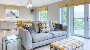 Ein modernes Wohnzimmer mit einem grauen Sofa, gemusterten Kissen, einem Glastisch und Blick ins Freie durch große Fenster.