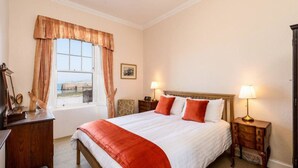 A hotel room with a large bed, two bedside tables with lamps, a wooden dresser, a window with curtains, and a view of the sea.