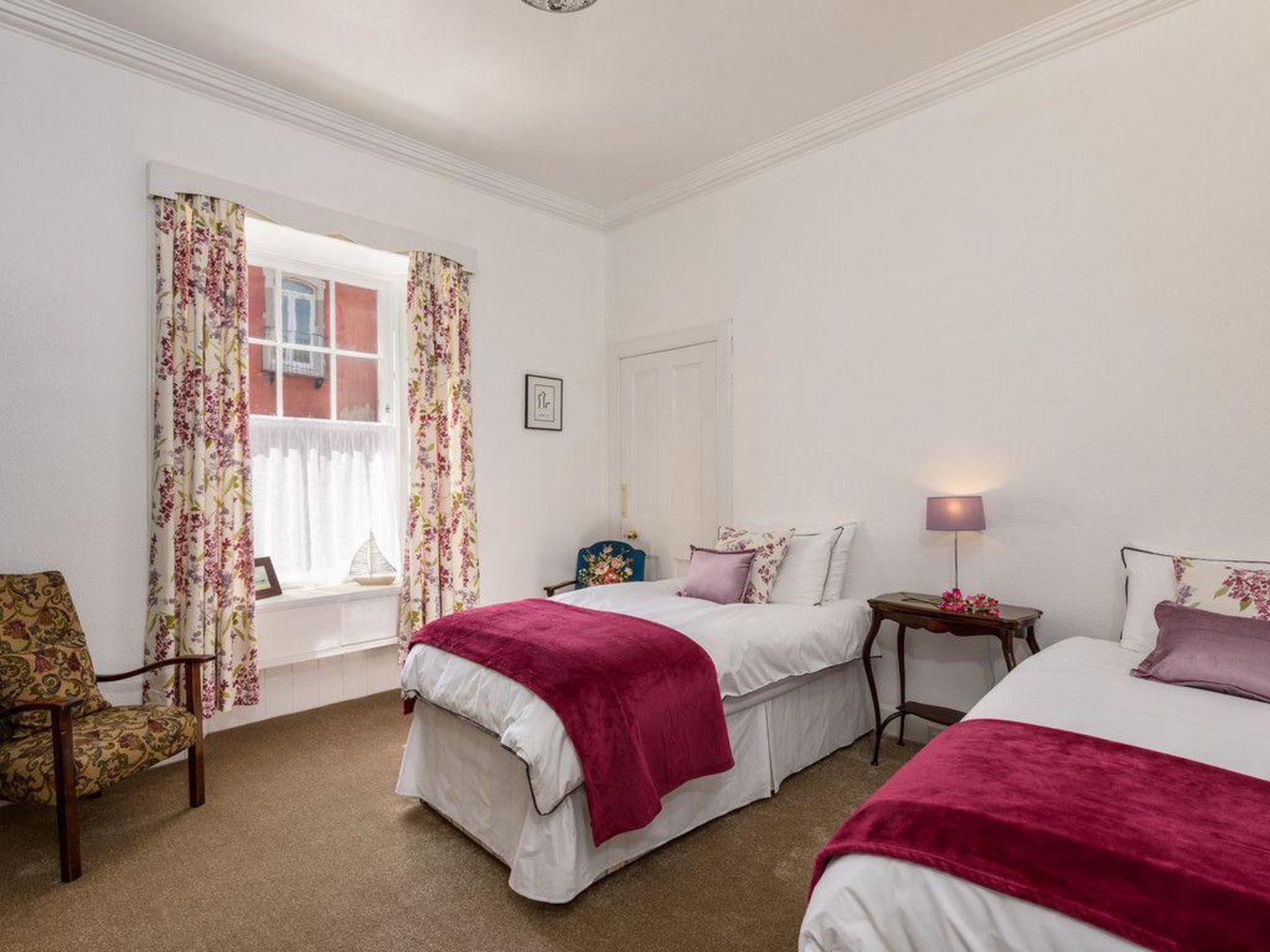 Une chambre d’hôtel avec deux lits, une fenêtre ornée de rideaux à motifs floraux, une chaise et une table de chevet avec une lampe.