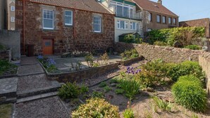 A stone house with a tiled roof, a front garden with various plants, and a stone wall.