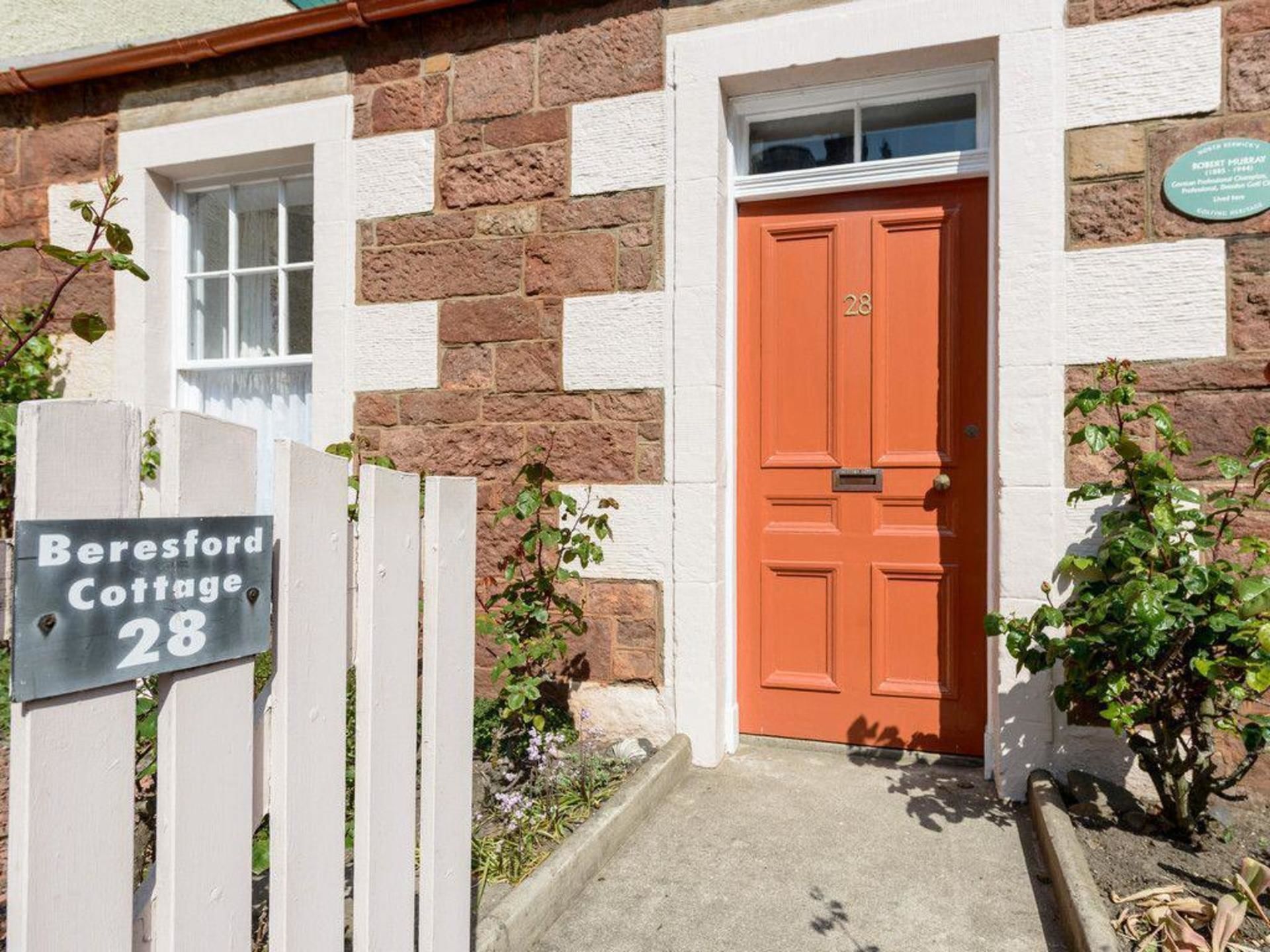 Une petite maison avec une porte rouge, une clôture blanche en piquets et une pancarte indiquant qu’il s’agit du numéro 28.