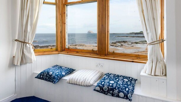 A room with a bay window overlooking a beach, featuring two pillows and a small bench. - Bass Rock View (North Berwick)