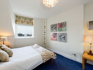 A bedroom with a bed, a window with a floral blind, a wall with four framed artworks, a bedside table with a lamp, and a blue carpet. - Bass Rock View (North Berwick)