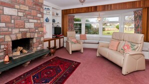 A cozy living room with a brick fireplace, a sofa set, a television, and a large window with curtains. - Greylag Cottage (Gullane)