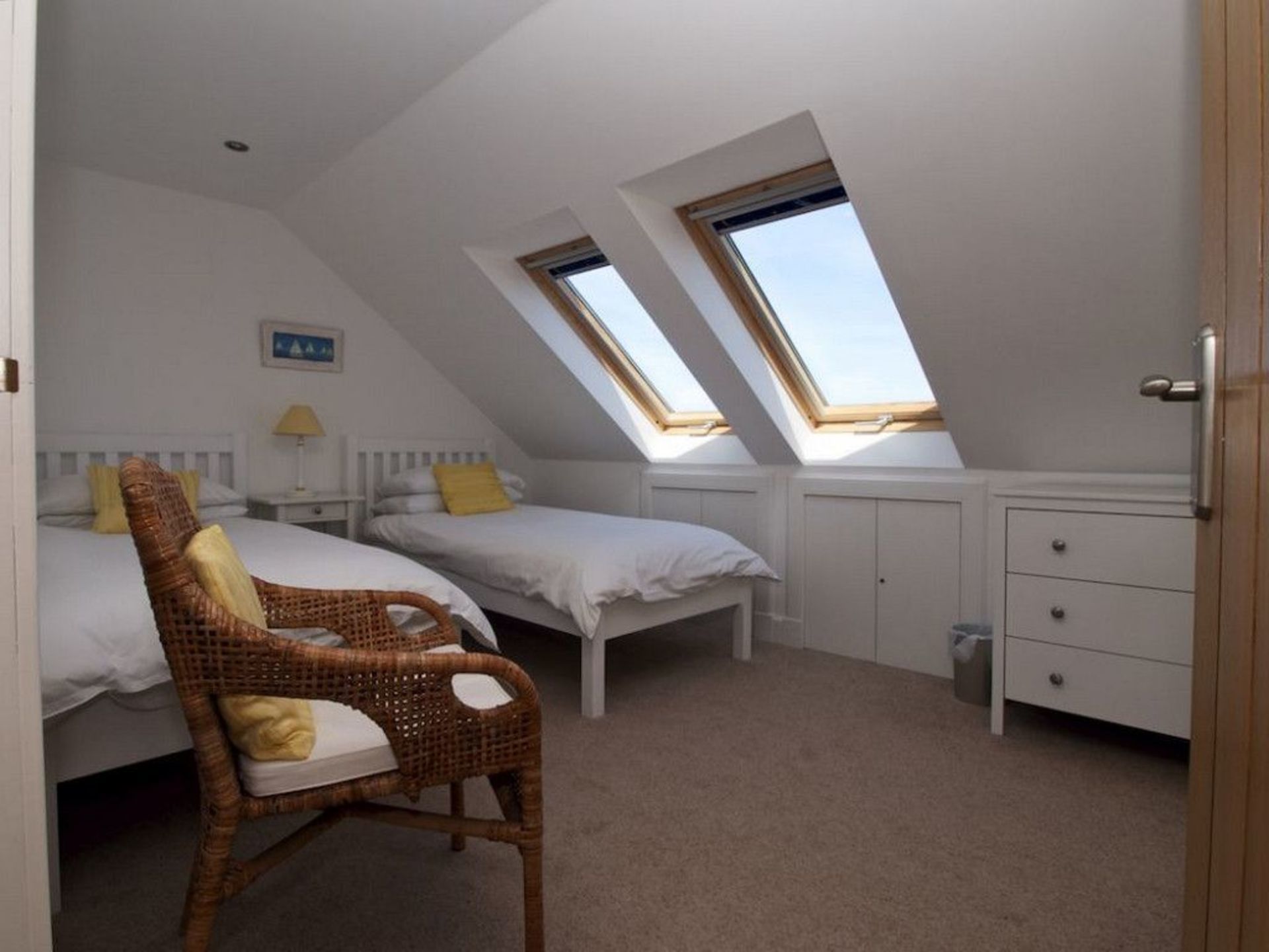A small, tidy room with two beds, a chair, and a desk. There are skylights allowing natural light.