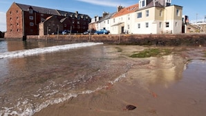 A coastal building with a brick facade and a white upper section, adjacent to a sandy beach with gentle waves. - Quayside Cottage (North Berwick)
