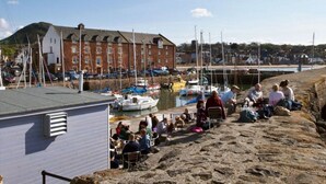 A coastal area with a stone wall, a building with a sloped roof, and several boats docked in the water. - Quayside Cottage (North Berwick)