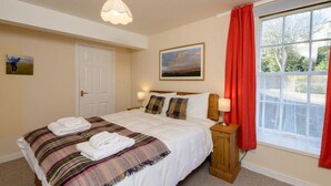 A neatly arranged bedroom with a bed, a bedside table with lamps, a door, a window with red curtains, and a painting on the wall. - Southcot (North Berwick)