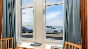 Una habitación con un ventanal grande, dos sillas, una pequeña mesa de madera con un libro y vistas al mar y un coche.