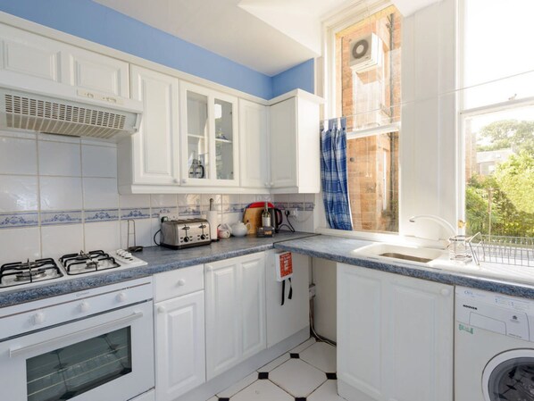 A kitchen with white cabinets, a gas stove, a built-in oven, a window with a blue curtain, and a washing machine.
