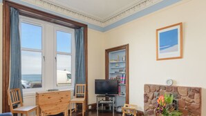 Una sala de estar con chimenea de piedra, televisor, estantería y vista al mar a través de amplios ventanales.