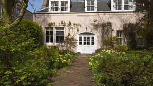 A traditional house with a brick pathway leading to a white door, surrounded by lush greenery and daffodils. - West Links View (North Berwick)