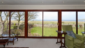 A room with a large sliding glass door leading to a balcony with a view of the sea. There is a green armchair and a small table on the balcony. - West Links View (North Berwick)
