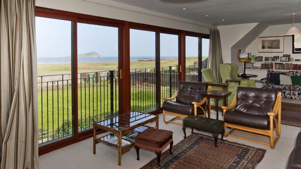 A spacious living room with large windows offering a view of the sea, a balcony with a railing, and a cozy seating area with leather chairs and a coffee table. - West Links View (North Berwick)