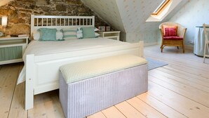 A bedroom with a sloped ceiling, a bed with a white headboard, a wicker bench, a chair, and a small table with a lamp. - Nungate (North Berwick)